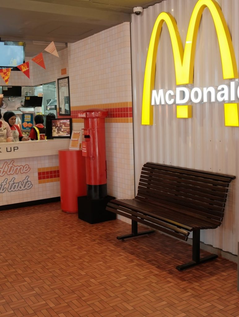 A retro-style McDonald’s interior featuring a large yellow McDonald’s logo, a wooden bench, a vintage red postbox, and a tiled counter area where staff are serving customers